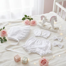 A white sissy baby accessories set arranged on a soft white blanket, including matching bonnet, panties, mittens, and two hair bows. A small white rocking horse toy, pink roses, wooden blocks, and warm string lights decorate the scene.