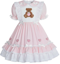 A pink and white adult baby girl's dress featuring a white bib front with an embroidered brown teddy bear holding a pink heart, short puff sleeves, and a tiered skirt decorated with pink hearts and white bows.