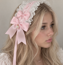 A blonde woman wearing a satin and lace headband decorated with a pink satin bow, a pearl accent, and long ribbon tails, photographed against a soft neutral background.