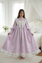 A woman wearing a lavender and white long-sleeved nightgown with lace details, standing barefoot in a bright bedroom.