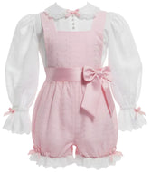 An adult baby girl's pink shortall romper with a matching pink waist bow, styled over a white long-sleeve blouse with ruffled cuffs.
